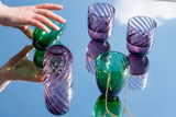 Violet Marika Tumblers on a mirror reflecting the sky, accompanied by gold jewellery and a moving hand 