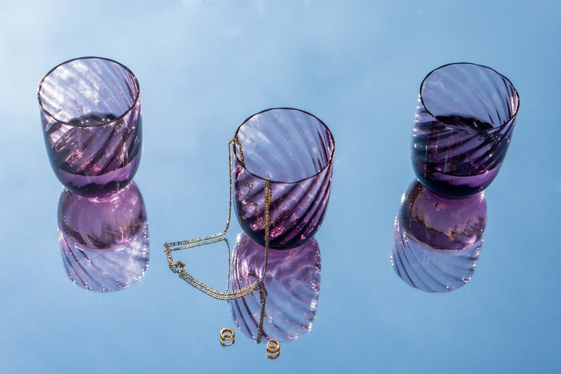 Violet Marika Tumblers on a mirror reflecting the sky and accompanied by gold jewellery 