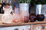 Violet Hobnail Tumblers on a wooden piece of furniture, surrounded by products from the Hobnail collection with wine bouiteilles and metals in the background
