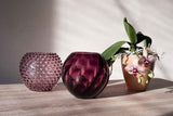 Violet Marika Vase on a wooden cabinet with a purple Hobnail vase on the left, a white and purple orchid on the right, and a white background with shadows